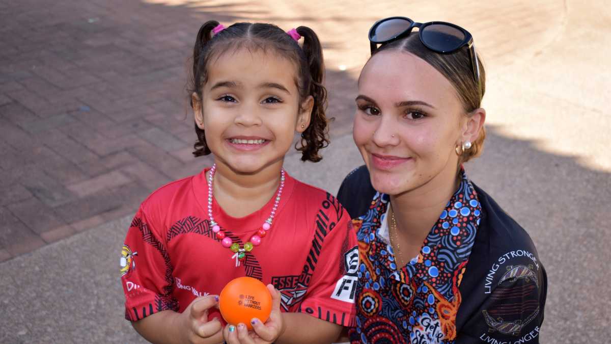 A young girl wearing a red top and holding an orange ball stands next to a woman in a polo with glasses on her head. Both are smiling at the camera.