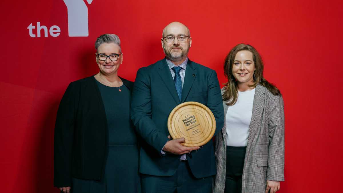 Mike, Fiona and another woman standing in front of a red background smiling at the camera. Mike is holding his award.