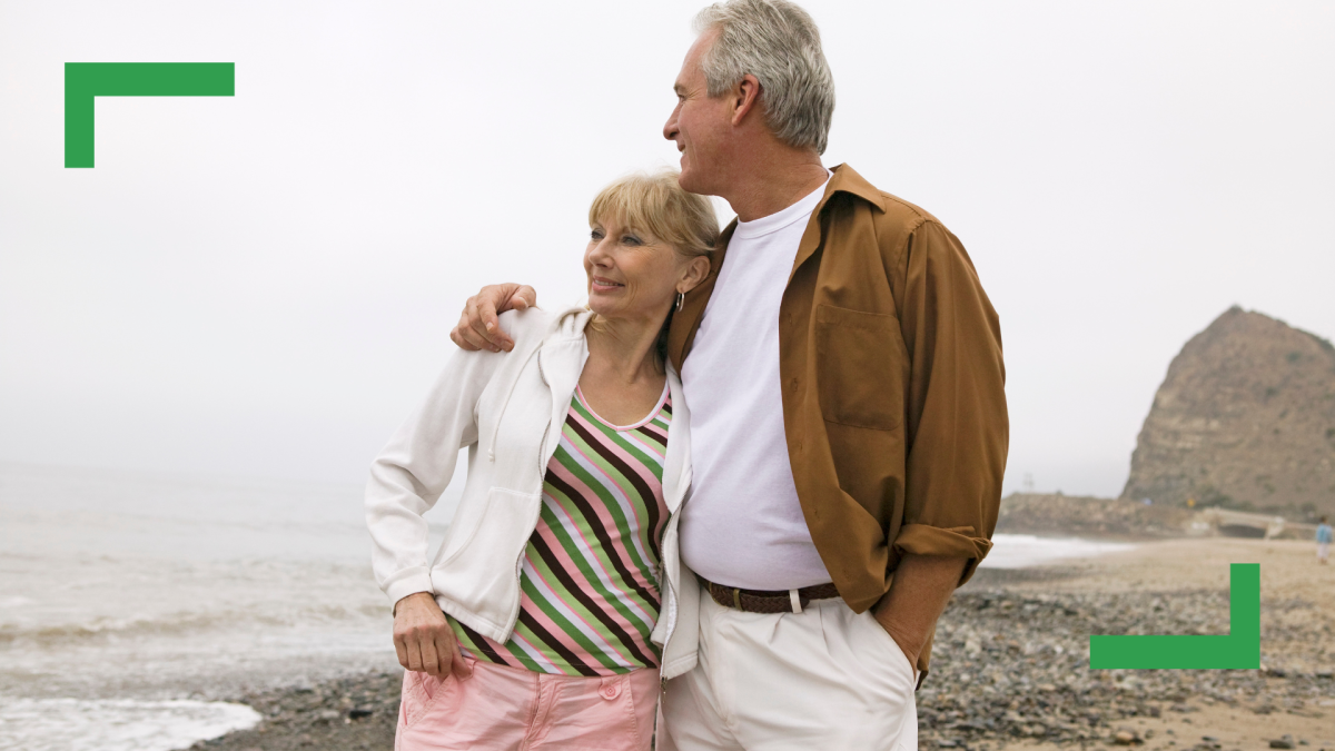 On a beach stands a middle-aged woman wearing pink pants, a striped shirt and a zip-up jacket standing with her husband who is wearing cream pants, a white shirt and a brown jacket. The man has his arm around the woman.