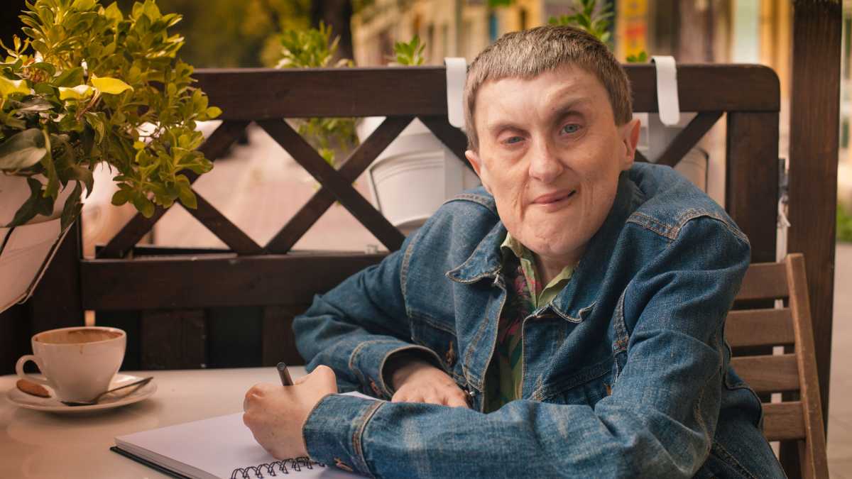 Man in a denim jacket writing in a notebook sitting at an outdoor café. He has a cup of coffee and plant on his table.