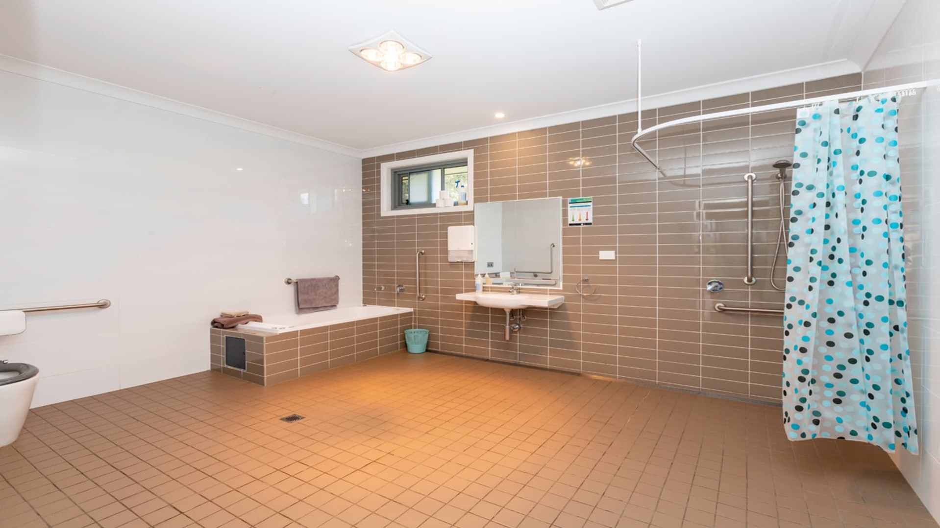 Accessible, spacious bathroom with brown tiles on floor and walls.  Stepless shower and blue shower curtain. small bathtub and basin with wheelchair access.