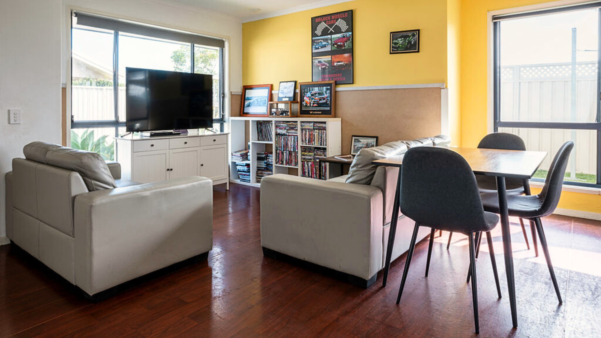 Lounge with two cream coloured couches, a small dining table, a large TV and a shelf.
