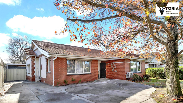 Single-storey brick house with tiled roof, front windows, and a tree casting shadows on the driveway under a partly cloudy sky.