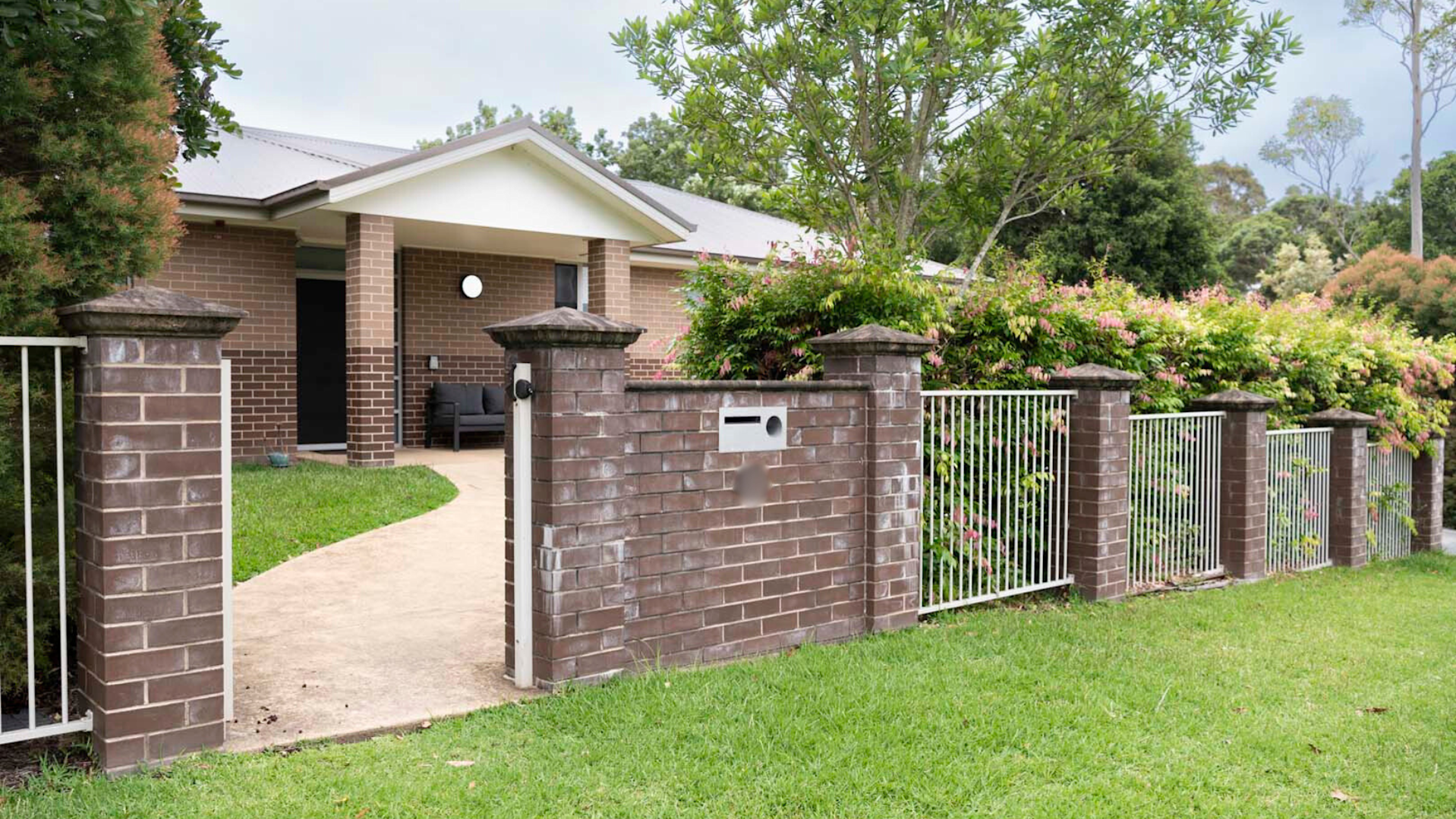 Front of the property, a brick fence with gate, there is a concrete path leading to the front door of the single story home.