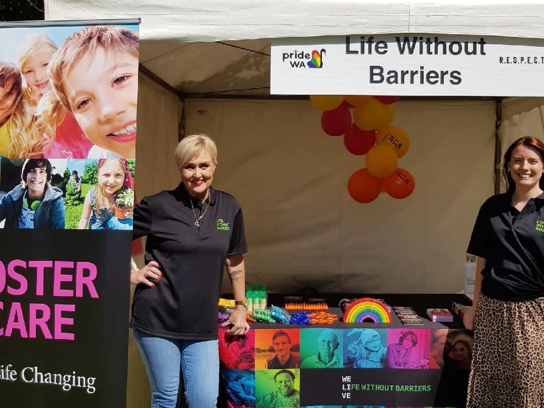 Sherry-Lee Vincent, Carer Engagement Officer for Western Australia, dressed in a black t-shirt with short blonde hair. she has rainbows painted on each cheek. she is standing in front of a Life Without Barriers tent with Sarah Broderick