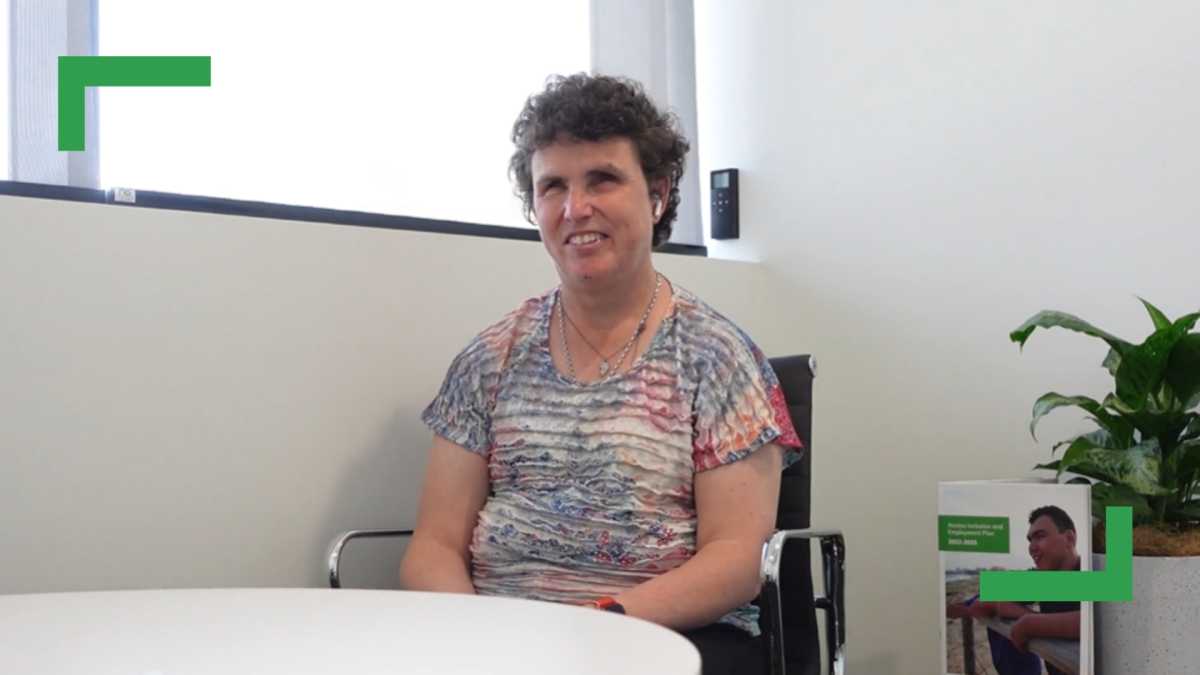 Emma is siting in an office room on a black chair with a round white table. She is smiling at the camera and wearing a pastel-coloured flower top. Behind her is a window and a plant.