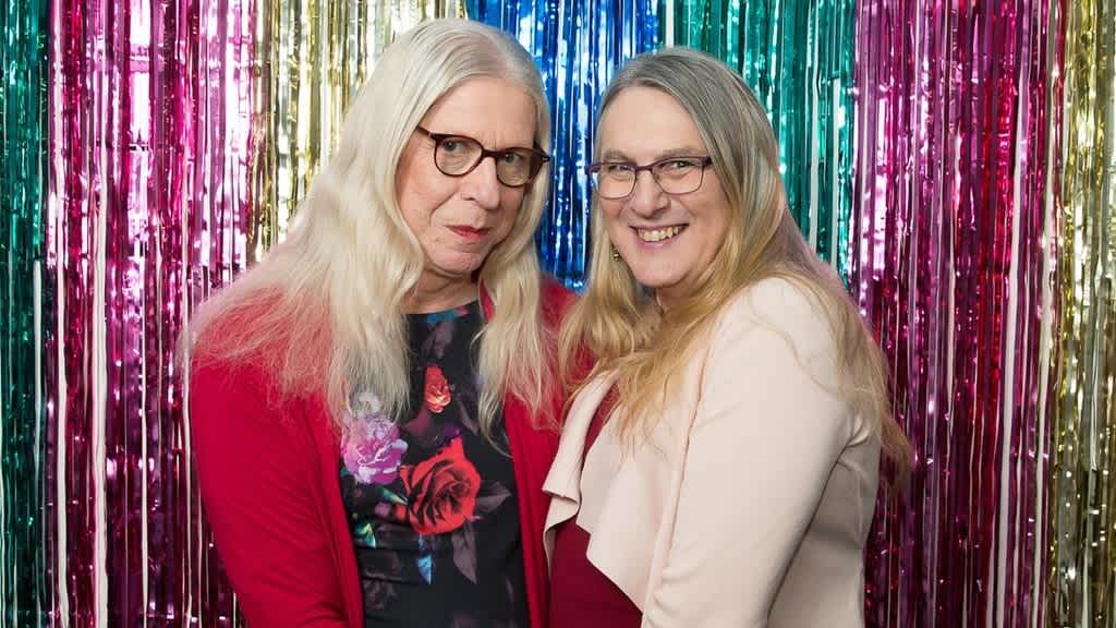 Gail and Lisette standing in front of rainbow streamers. Both are wearing glasses and have long hair.