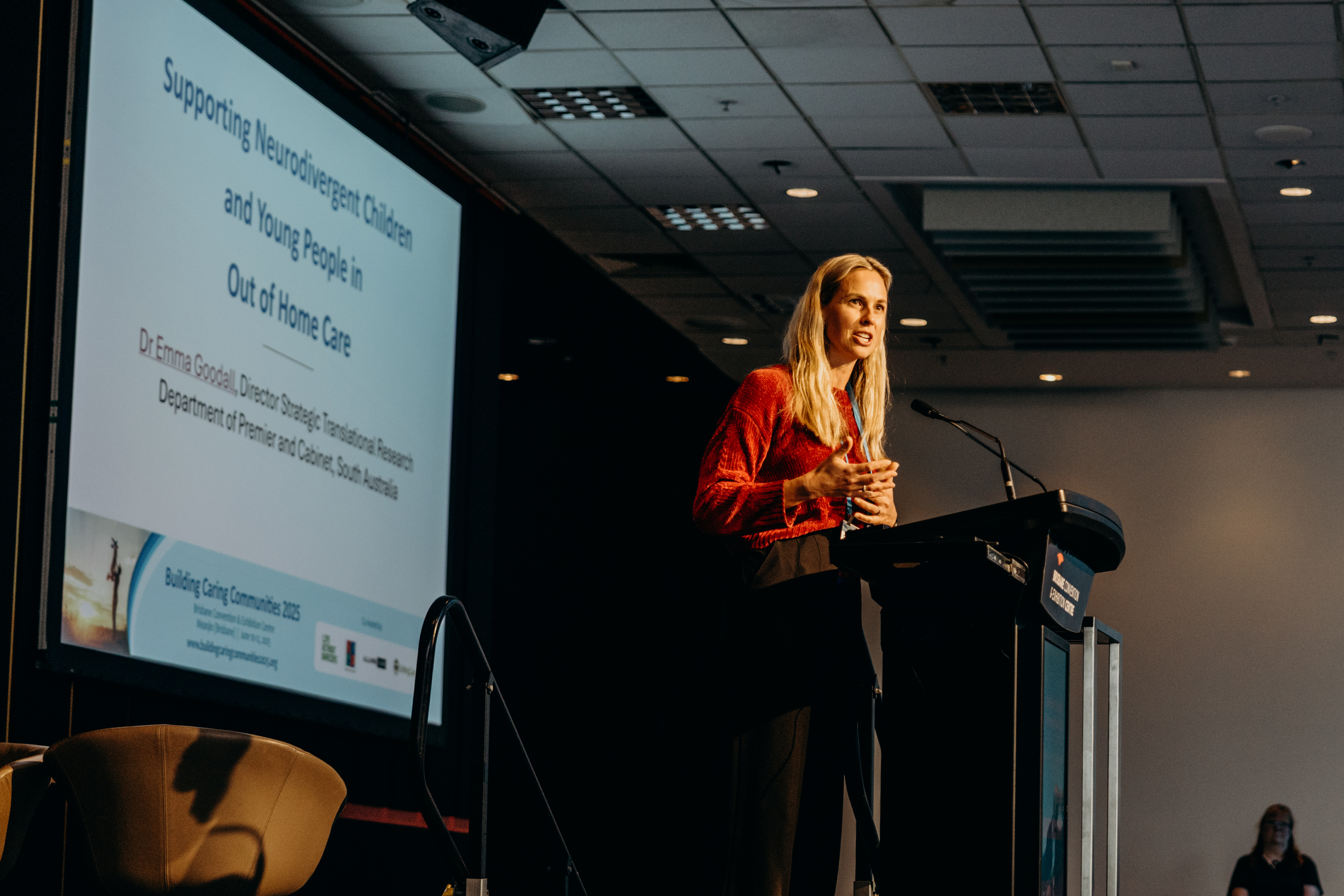 Courtney is one stage speaking. She is wearing a red shirt and standing in front of a podium.