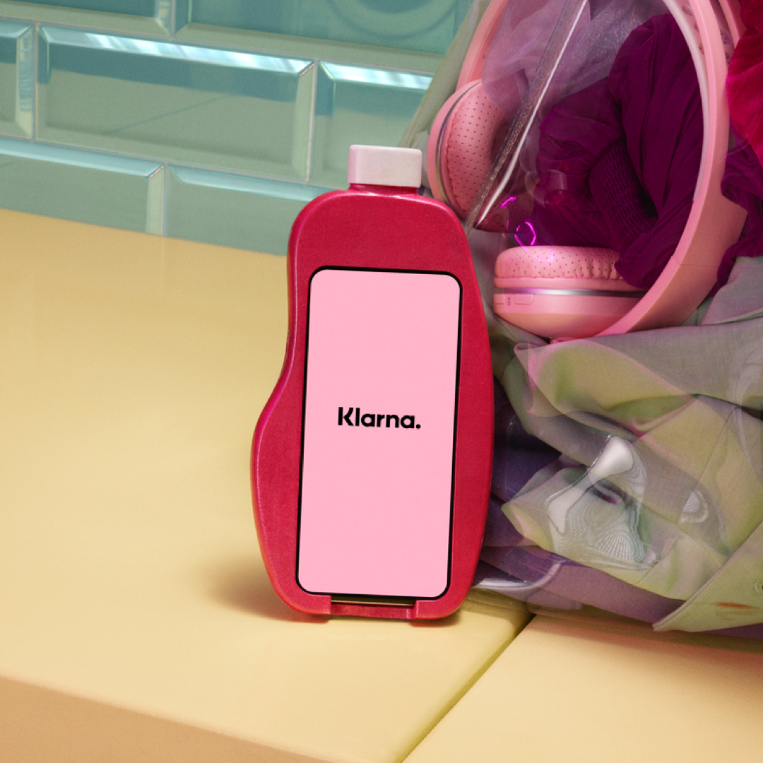 (Instagram) Un téléphone affichant le logo Klarna dans un étui en forme de bouteille, avec des écouteurs roses en arrière-plan