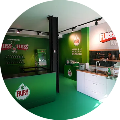Interior de una cocina con una encimera blanca, productos de limpieza y paredes verdes con logotipos de Fairy.