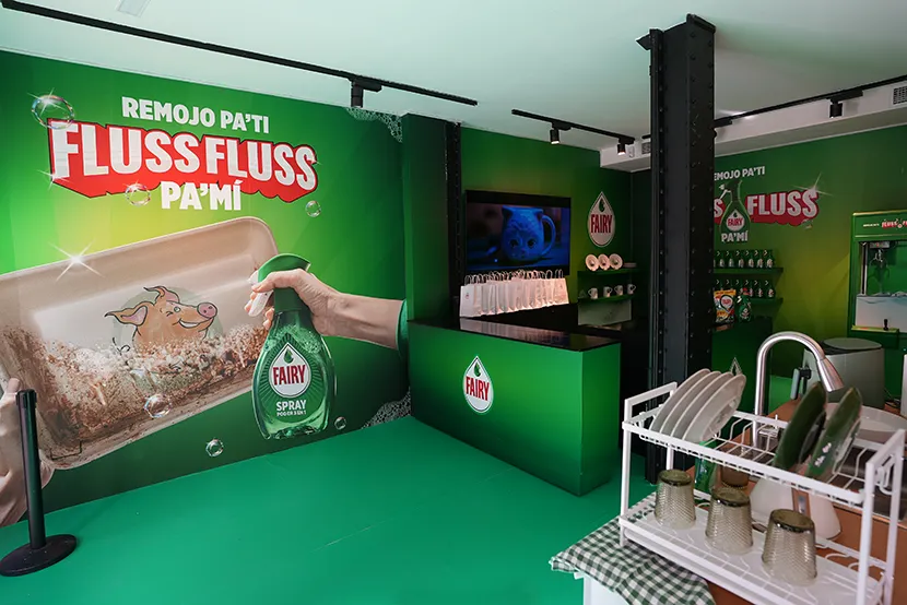 Pared verde con texto REMOJO PA'TI FLUSSFLUSS PA'MI, botella de Fairy en mano, mesa negra con platos, estantería con productos y pantalla.