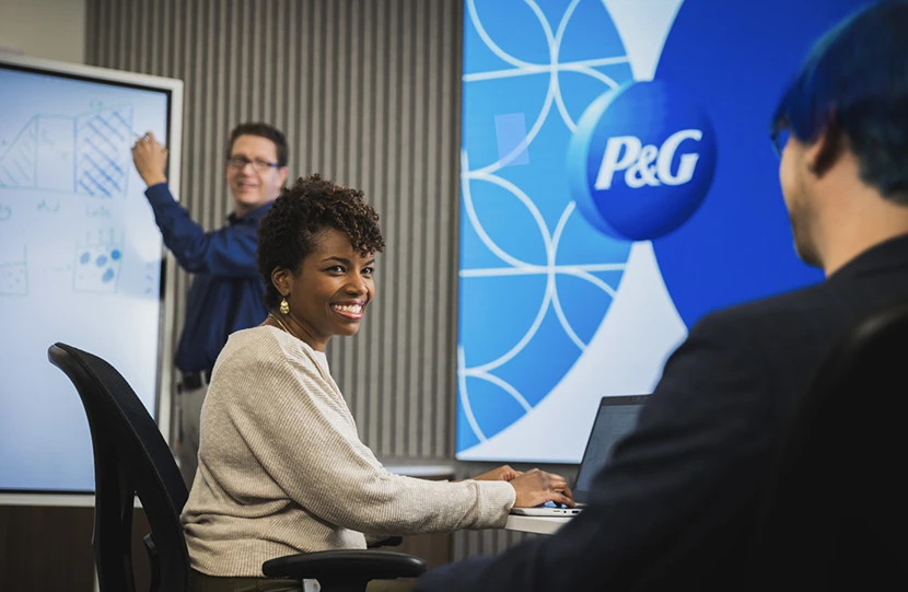 Una empleada y dos empleados de P&G colaborando en el espacio de trabajo de P&G. La empleada y el logotipo de P&G están en el centro de la imagen y los empleados están a cada lado.