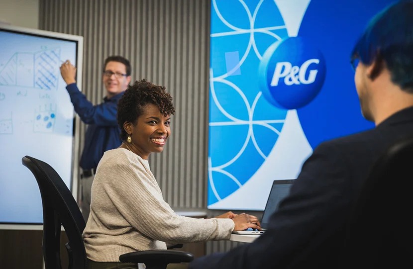 Una empleada y dos empleados de P&G colaborando en el espacio de trabajo de P&G. La empleada y el logotipo de P&G están en el centro de la imagen y los empleados están a cada lado.
