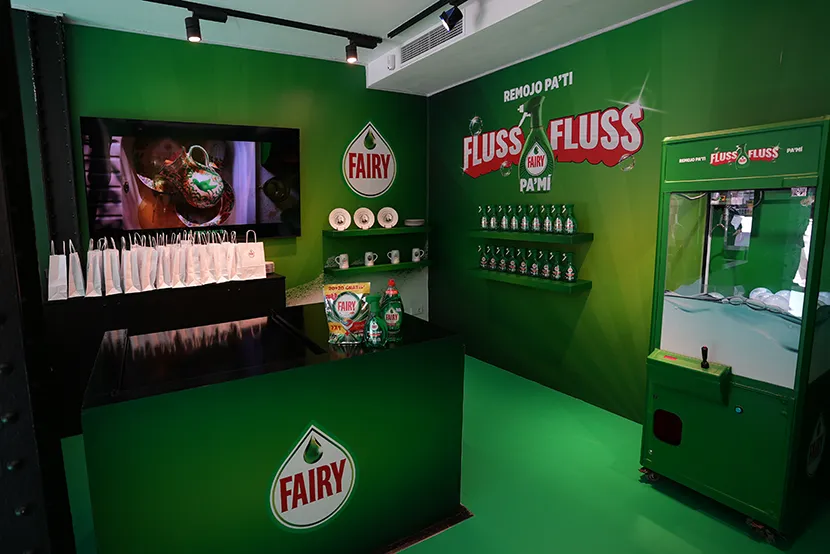 Tienda con pared verde, mesa negra, productos de Fairy y pantalla mostrando un video sobre limpieza.