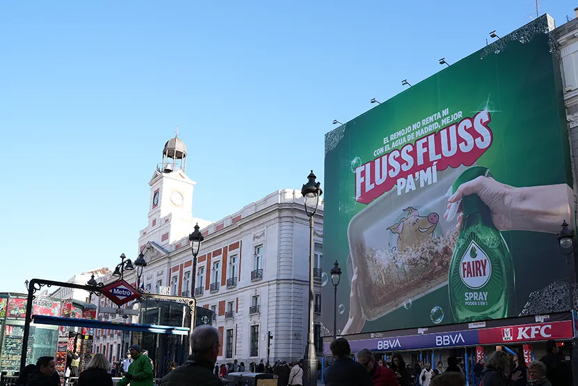 Edificio con torre y reloj, cartel grande de Flussfluss, personas y metro en la plaza bajo un cielo azul claro.