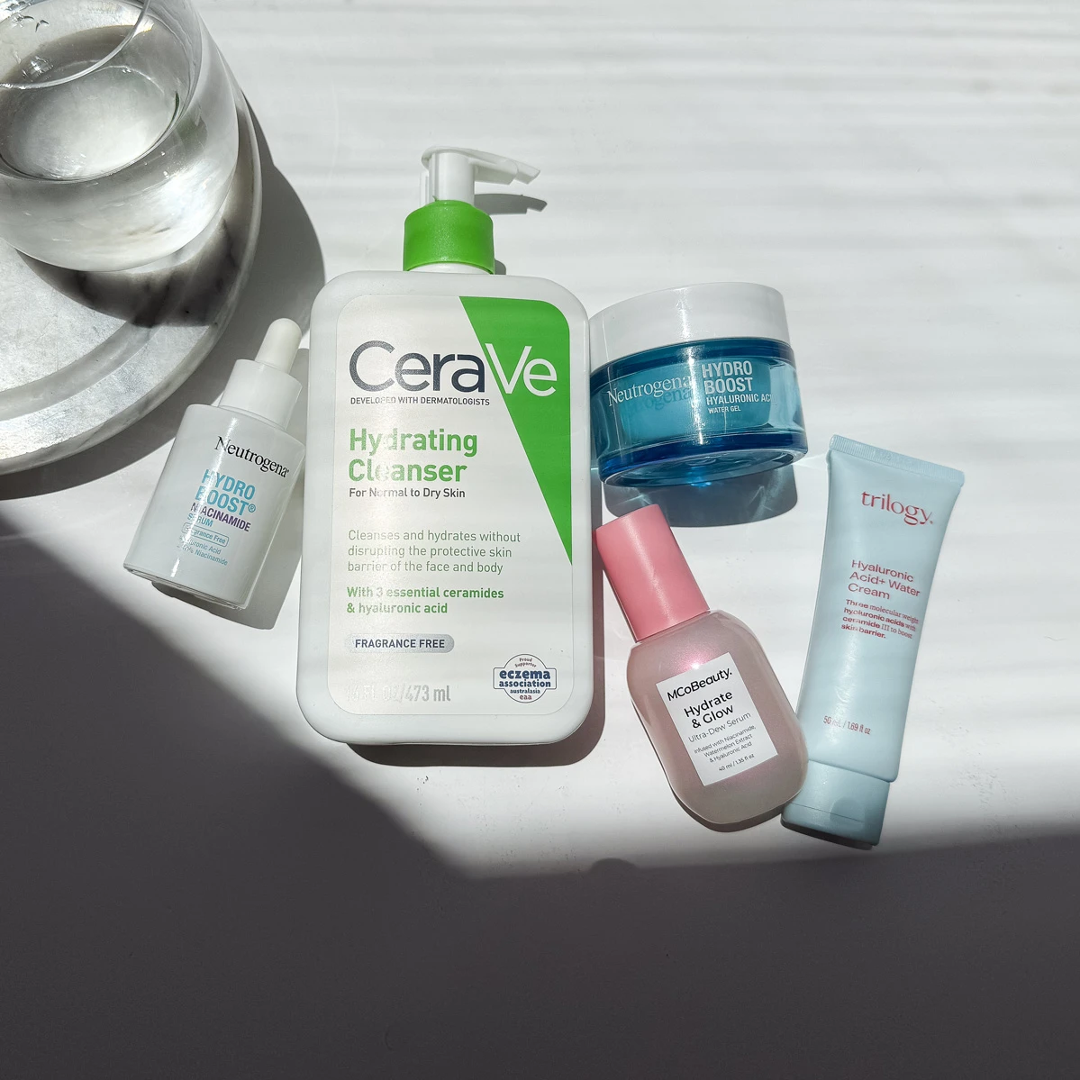 Collection of skincare products including CeraVe cleanser, Neutrogena Hydro Boost, and Trilogy cream arranged on white surface with shadows