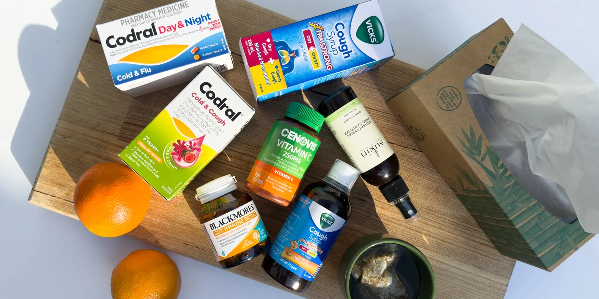 Cold and flu remedies on a wooden board, including Codral, Vicks cough syrup, Cenovis Vitamin C, Blackmores, hand sanitiser, tissues, and oranges.