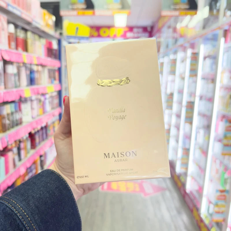 Hand holding Maison Asrar Vanilla Voyage 100ml Eau de Parfum box in a store aisle with shelves in the background.