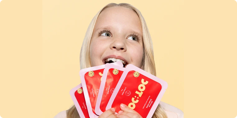 A young blonde girl smiles and looks up while holding several red and pink Loc:Loc product packets against a pale yellow background.
