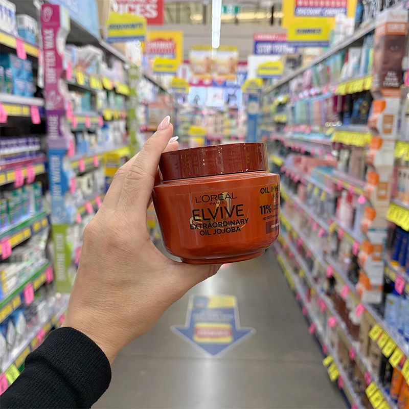 A hand holds a red jar of L'Oreal Paris Elvive Extraordinary Oil Jojoba hair mask in a retail store aisle.