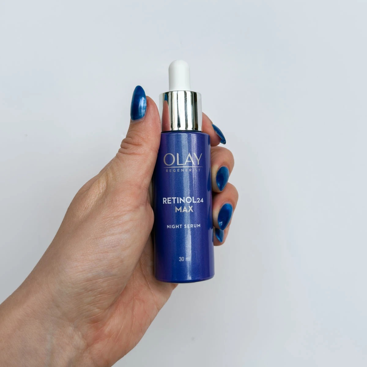 Hand with blue nail polish holding a purple bottle of Olay Regenerist Retinol24 Max Night Serum against white background
