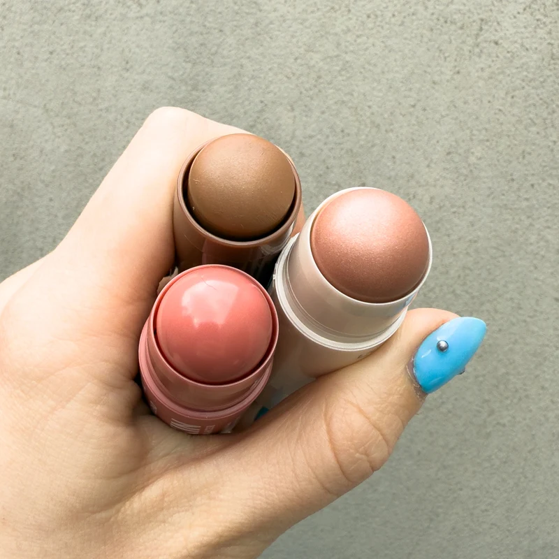 Hand holding three makeup blush sticks in pink, bronze, and rose gold shades, with a blue manicured thumbnail visible.