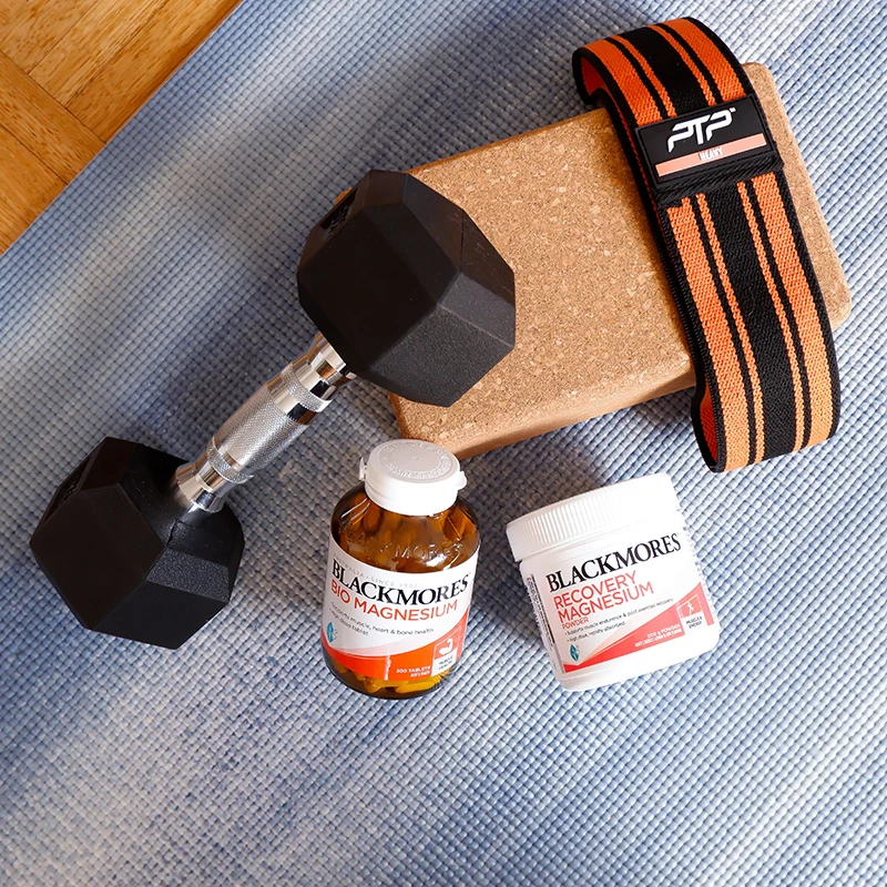 Workout equipment including a dumbbell, weightlifting belt, yoga block, and Blackmores magnesium supplement bottles on exercise mat.