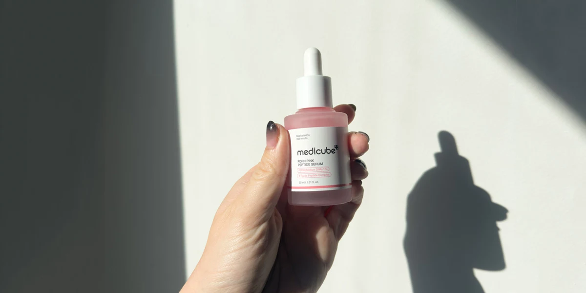 A hand holding a Medicube PDRN Pink Peptide Serum bottle with a white dropper cap against a sunlit white wall.