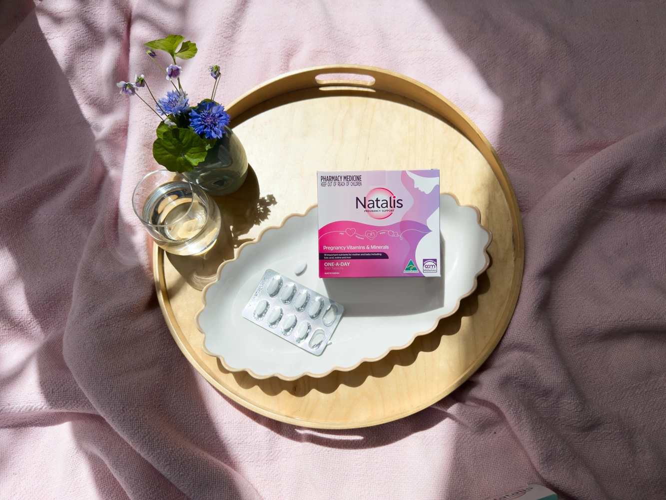 Natalis pregnancy vitamins box and blister pack on gold tray with blue cornflowers in vase and glass of water on pink fabric