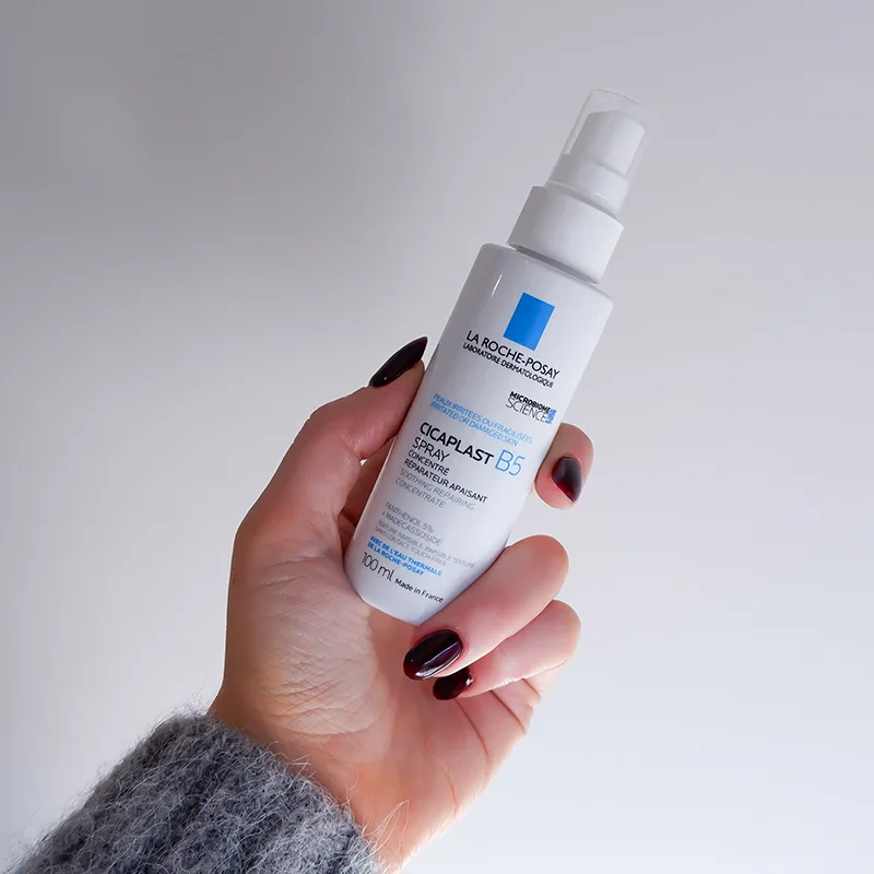 Hand with dark nail polish holding a white La Roche-Posay Cicaplast B5 spray bottle against a light background.