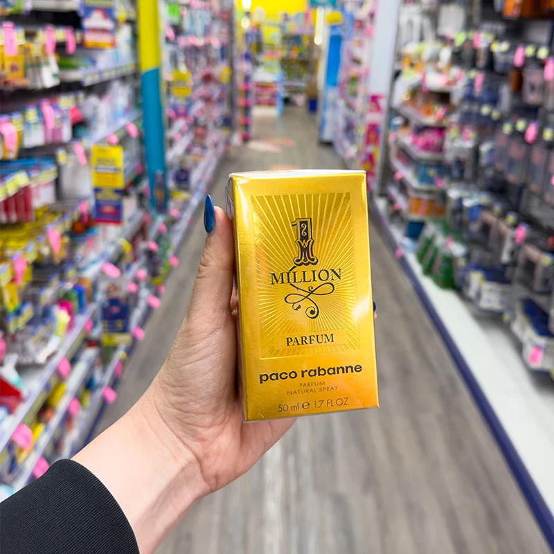 Hand holding gold Paco Rabanne 1 Million perfume box in retail store aisle with colorful product displays in background