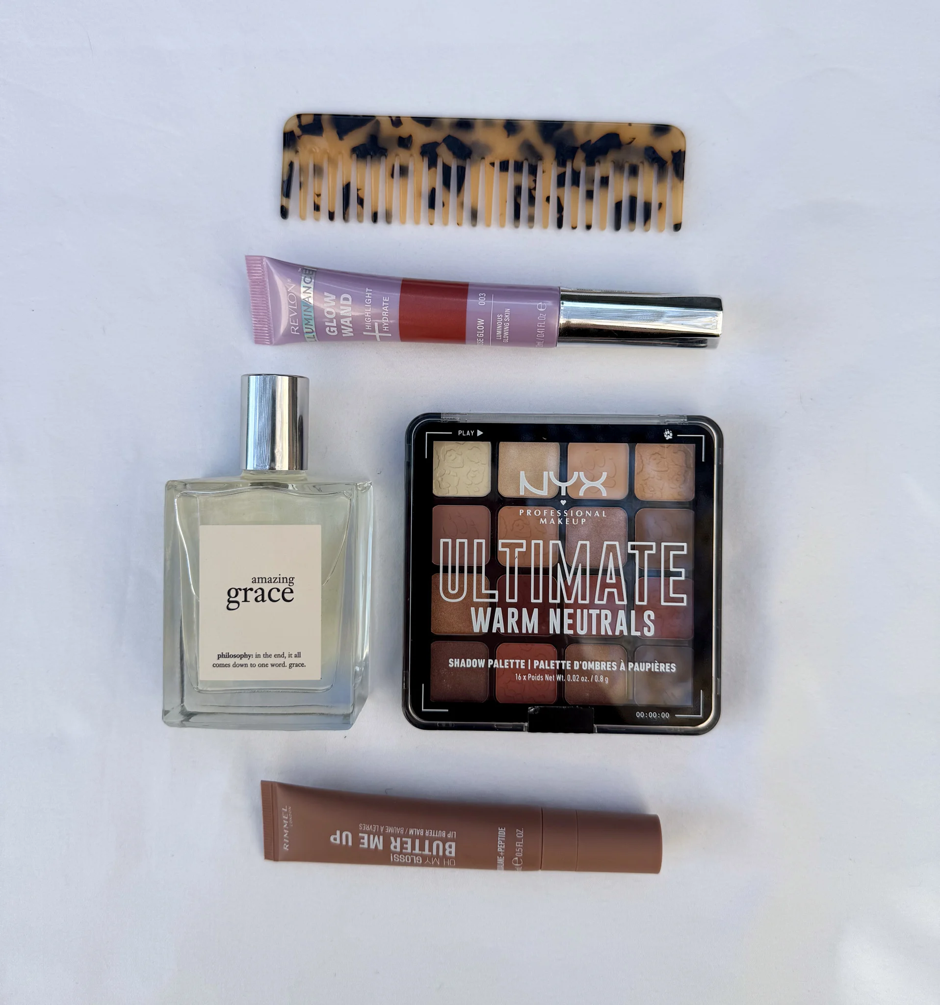 Beauty products flatlay featuring NYX eyeshadow palette, Amazing Grace perfume, lip products, and a tortoiseshell comb on white background.