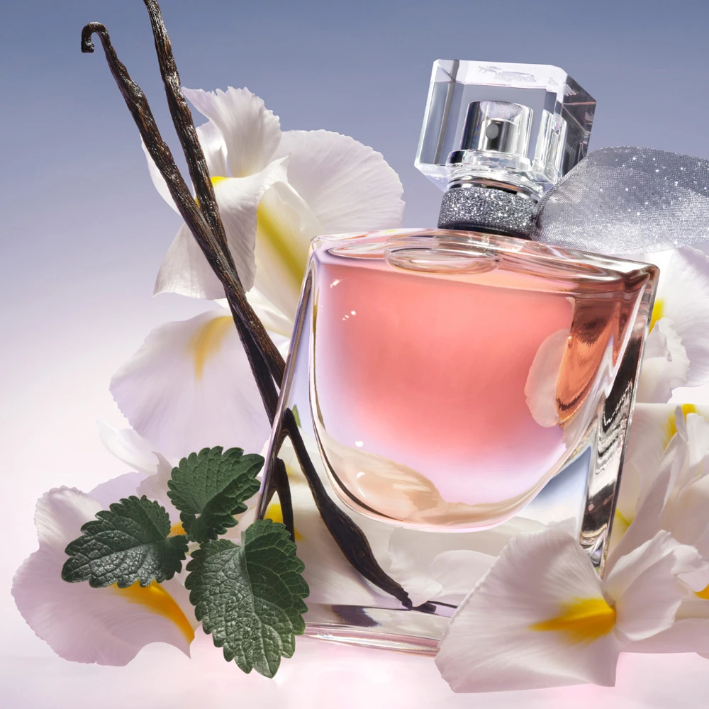 Pink Lancome perfume bottle with vanilla pods surrounded by white daffodils and green leaves against a light background