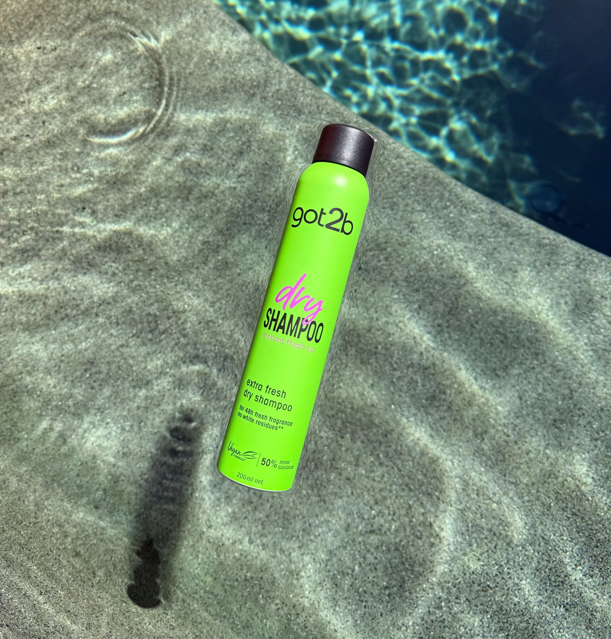 Bright green Got2b dry shampoo can on concrete surface near pool water with rippling reflections.