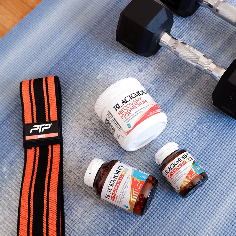 Workout equipment including a dumbbell and orange weightlifting strap alongside Blackmores supplement bottles on a blue yoga mat.