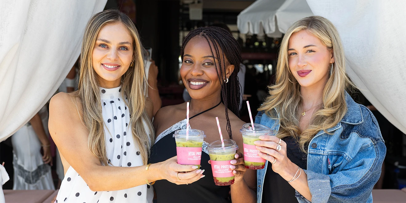 Three smiling friends enjoying colorful smoothies outdoors, wearing casual summer outfits including denim and polka dots.
