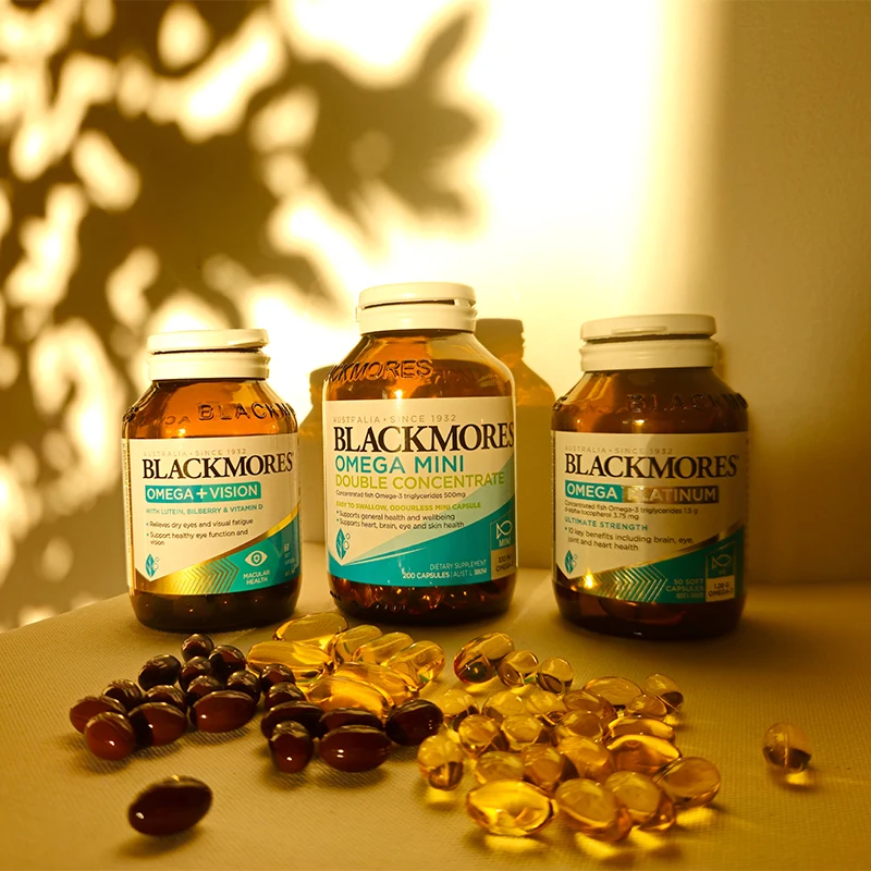Three Blackmores omega supplement bottles with scattered gel capsules in front, lit by warm golden light with leaf shadows on wall.