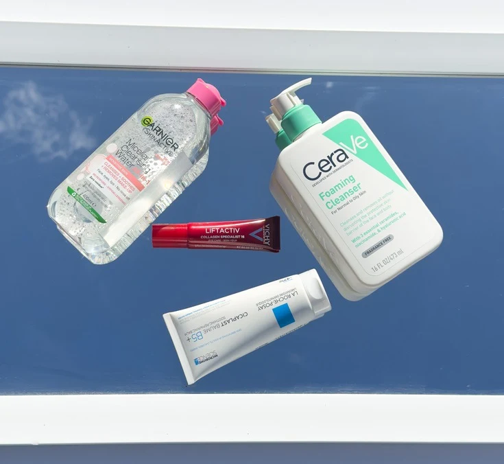 Four skincare products on a blue surface: Garnier Micellar Water, CeraVe Foaming Cleanser, Vichy Liftactiv serum, and La Roche-Posay Cicaplast Baume B5.