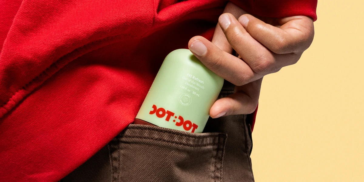 A hand places a light green Dot:Dot deodorant stick into a brown pants pocket, wearing a red hoodie, against a cream background.