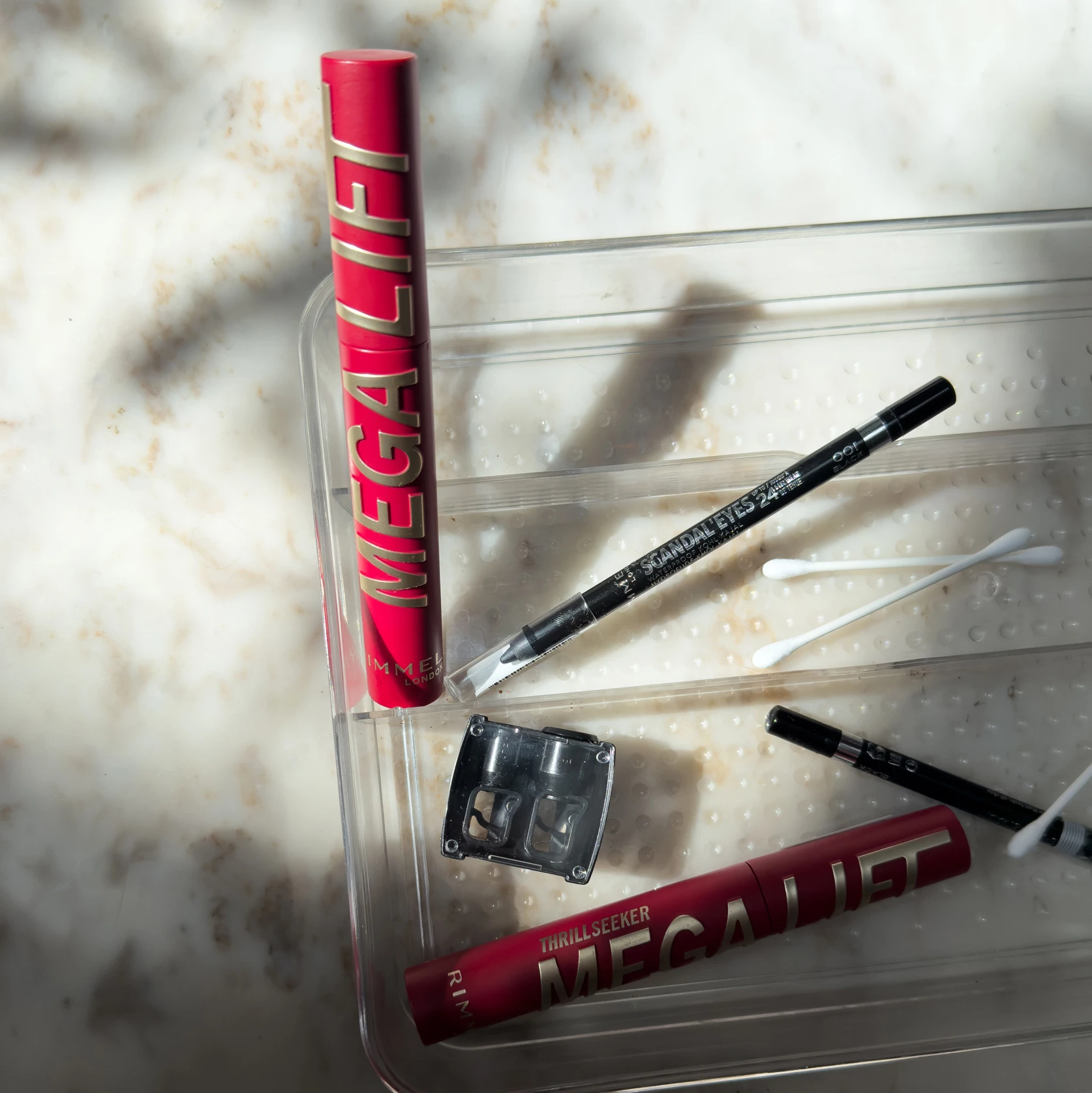Red Rimmel mascara tube and black eyeliner pencils scattered on clear plastic surface with soft shadowy lighting