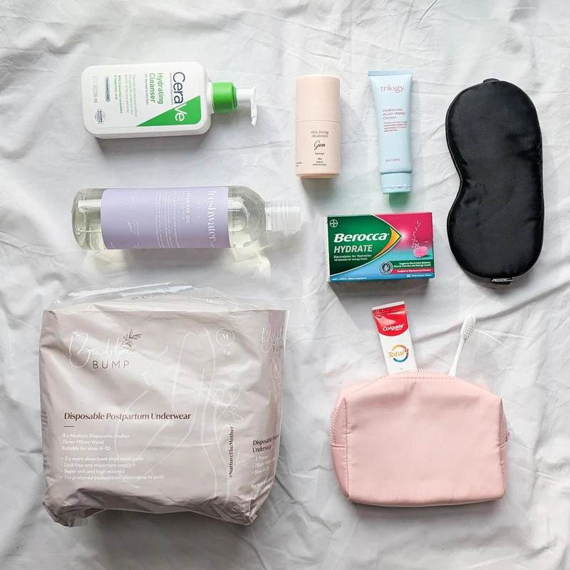 Postpartum essentials flatlay including CeraVe cleanser, Trilogy cream, Berocca, sleep mask, Colgate, pink cosmetic bag, and disposable underwear.