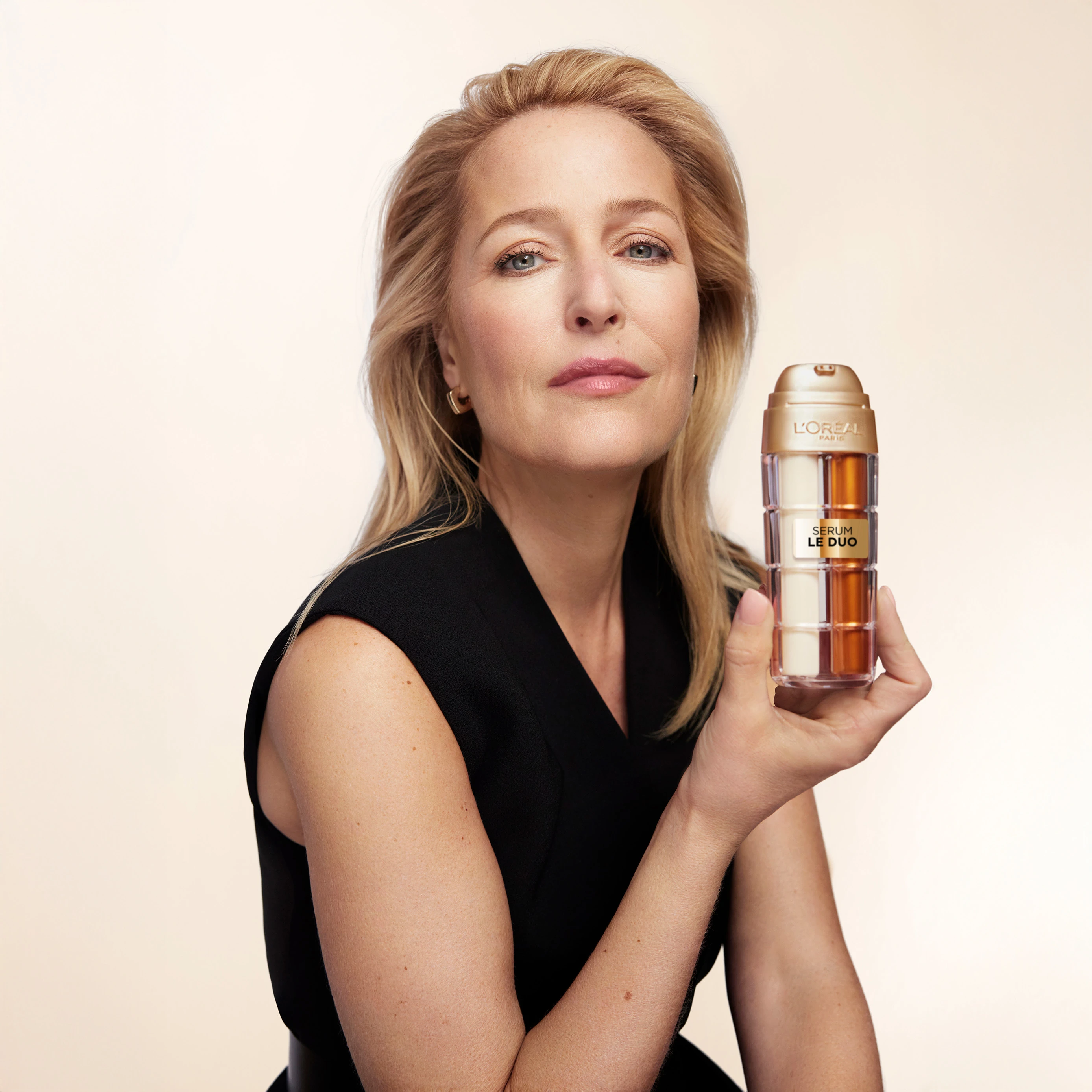 Woman in black sleeveless top holding L'Oreal Le Duo serum bottle against light background, showcasing beauty product.