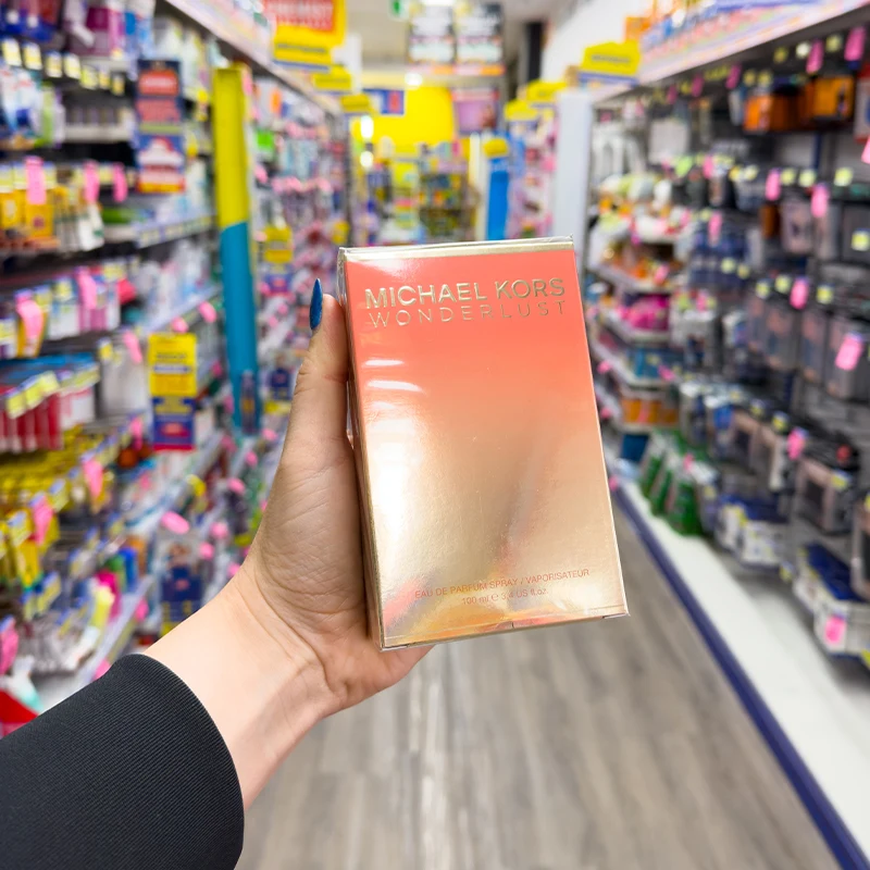 Hand holding pink Michael Kors Wonderlust perfume box in a retail store aisle with colorful product displays in background