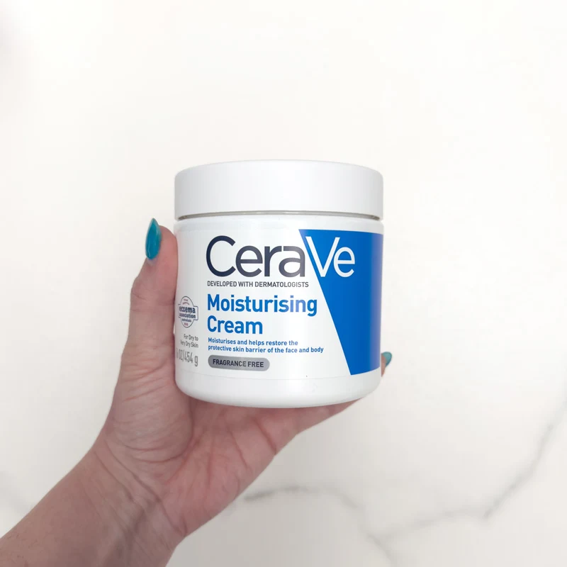 A hand with teal nails holds a white and blue tub of CeraVe Moisturising Cream, fragrance-free, against a white background.