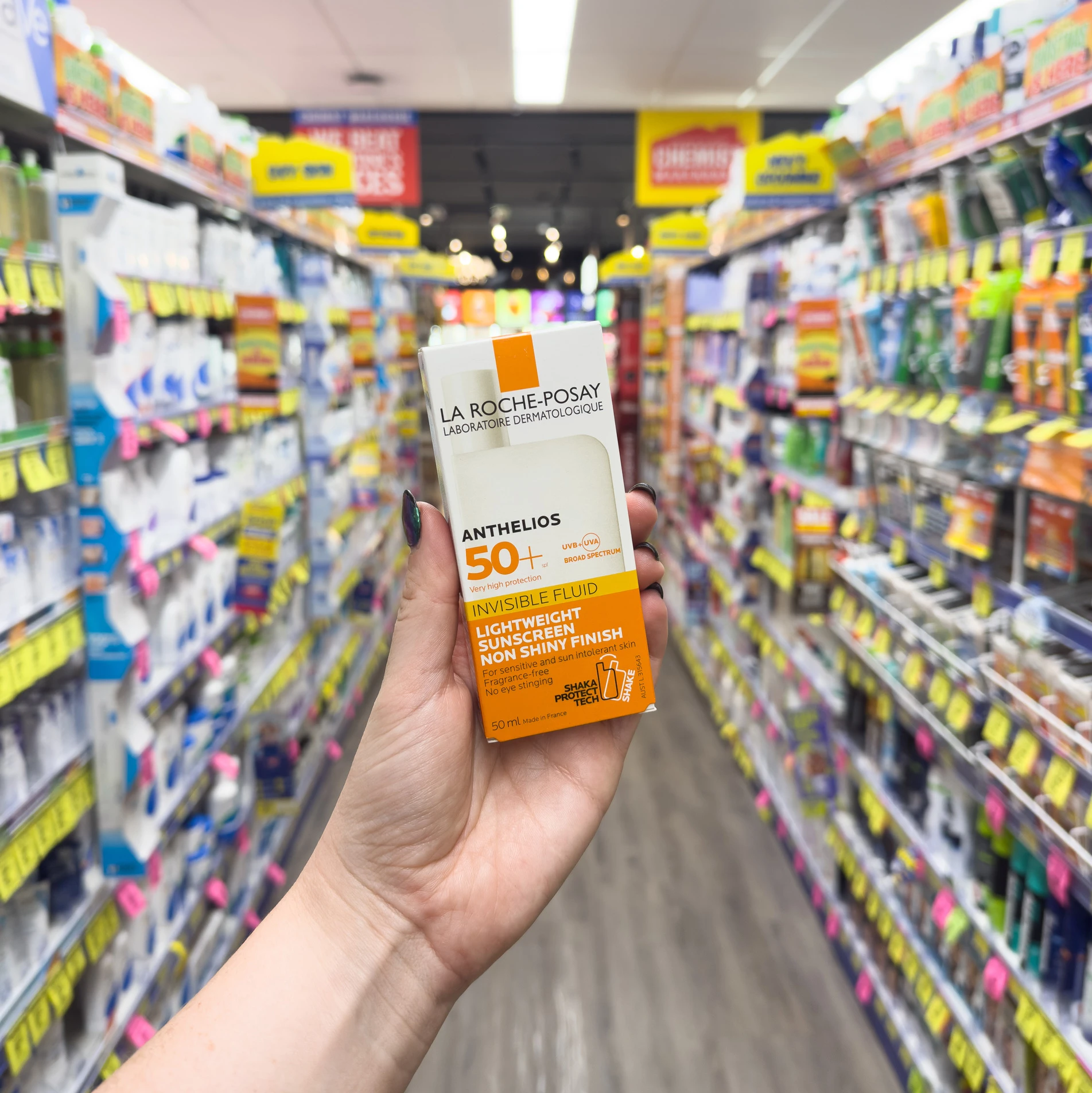 Hand holding La Roche-Posay Anthelios SPF 50+ sunscreen in a drugstore aisle with shelves of personal care products in background