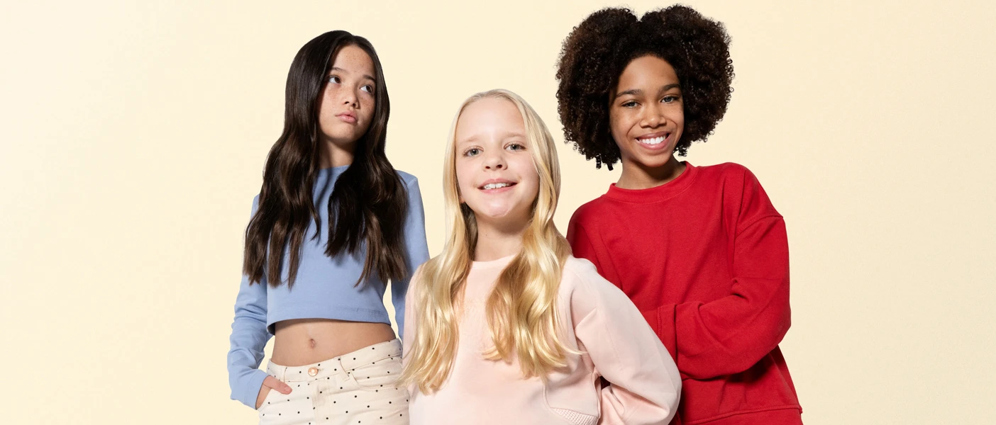 Three diverse girls posing together, wearing blue crop top, pink sweatshirt, and red sweatshirt against a cream background.