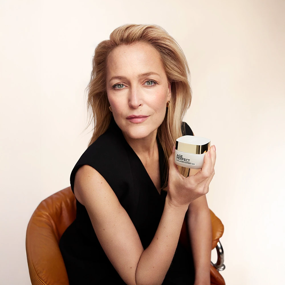Woman in black sleeveless top holding skincare product jar, seated on brown leather chair against light background.