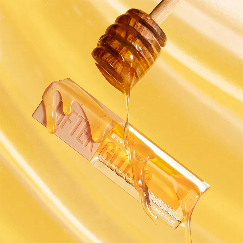 Golden honey drips from a wooden dipper onto a Maybelline Fit Me foundation bottle against a warm yellow background.