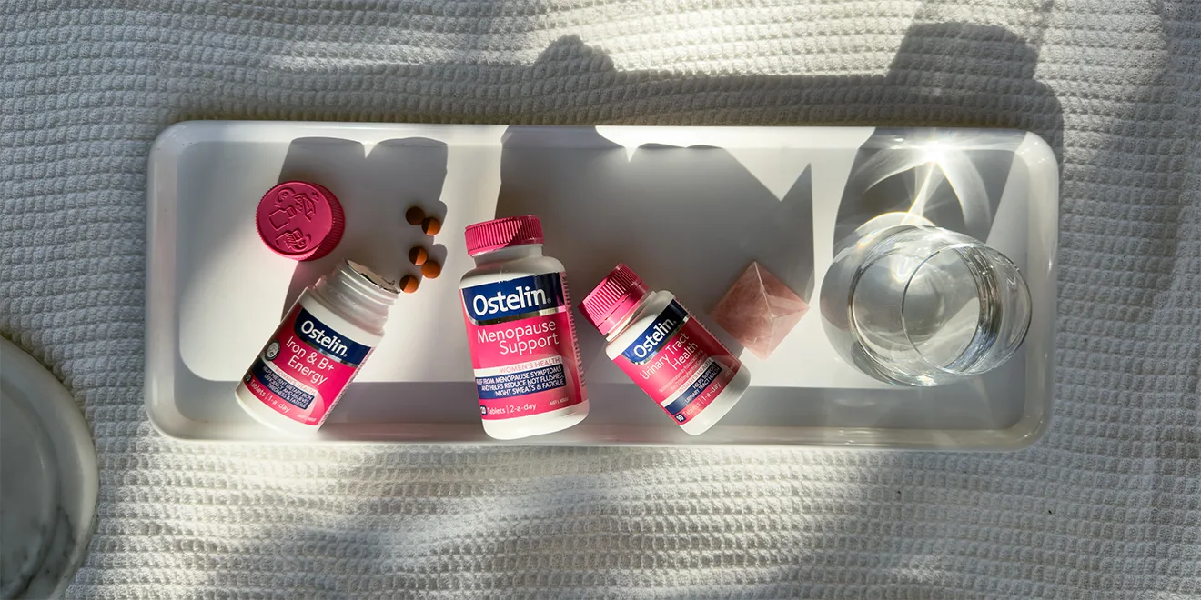 Three Ostelin vitamin bottles with pink caps on white tray with glass of water, showing menopause and energy supplement products.