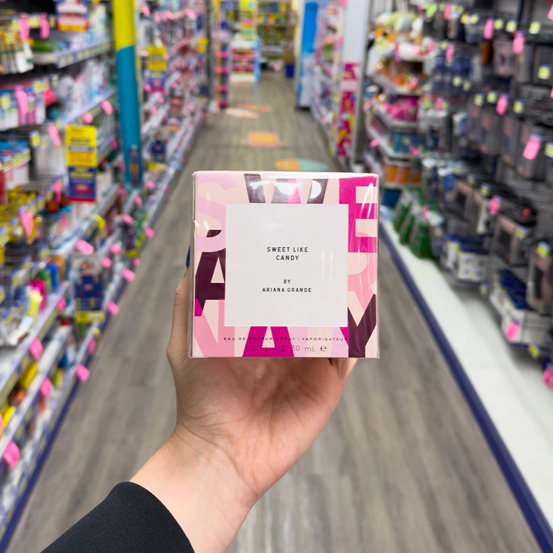 Hand holding Sweet Like Candy perfume box by Ariana Grande in a store aisle with blurred product shelves in background